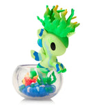 The tokidoki (IT) Neon Reef Mermicorno Blind Box features a green and blue seahorse-like toy with leafy hair in a clear bowl of blue pebbles and a plastic orange fish—an eye-catching addition to any collection.