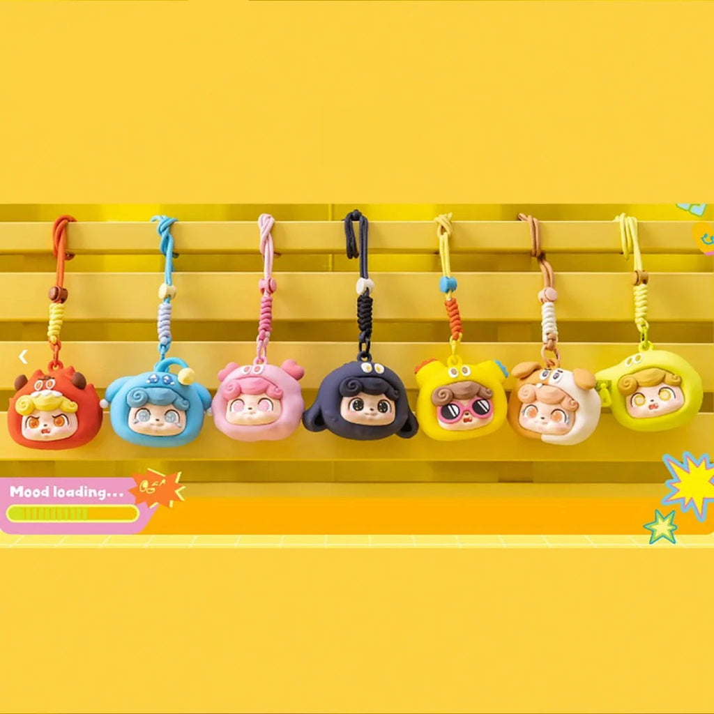 Seven playful cartoon keychains, including the Jotoys Q.Kid Emotional Monster Headphone Bag and animal hat designs, hang on a yellow rack against a yellow background, with a