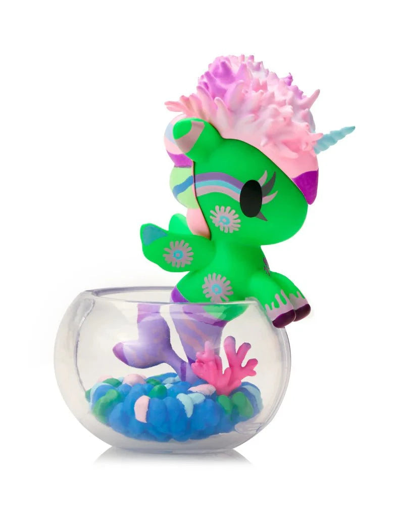 The tokidoki (IT) Neon Reef Mermicorno Blind Box features a colorful unicorn figurine with coral and shell accents, seated in a clear bowl filled with blue, green, and purple pebbles that glow under blacklight.