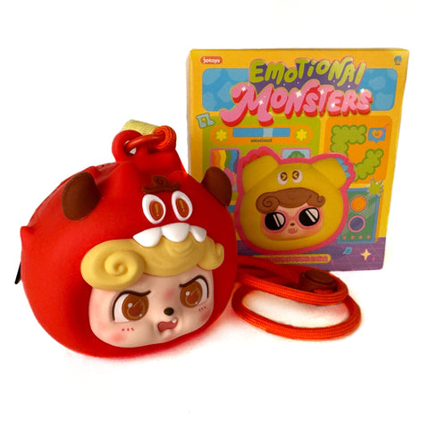 A round red Q.Kid Emotional Monster Headphone Bag by Jotoys, featuring a cartoon monster face with horns and yellow hair, sits before a colorful "Emotional Monsters" box. An orange strap makes it practical and fun to carry.