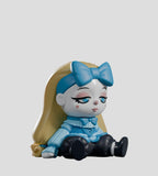 A vinyl doll from Kaleidos Creative’s A Mad Tea Party Blind Box, with long blonde hair, a big blue bow, blue sweater, black pants, and blue shoes sits on the ground with a neutral expression.