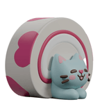 A blue cartoon cat with pink cheeks and brown whiskers sits in front of a roll cake, inspired by the Bake Me A Cat Blind Box figures from Kaleidos Creative.