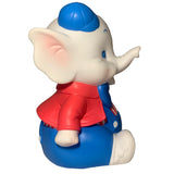 A side view of the Carnival Stripes Baby Elephant by Bitter Squeaks (US), a cartoon figurine in a blue cap, red jacket, and blue overalls, sits and points forward like a plush toy.