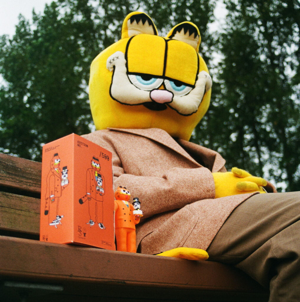A person in a large Garfield costume sits on a bench next to the Gangster Doodles GARF Vinyl Toy & Cassette Tape by Gangster Doodles and its box, with trees visible in the background.