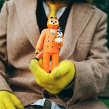 A person in yellow gloves and a brown suit holds the Gangster Doodles GARF Vinyl Toy & Cassette Tape by Gangster Doodles, featuring GARF in an orange suit with a small vinyl figure in its left arm.