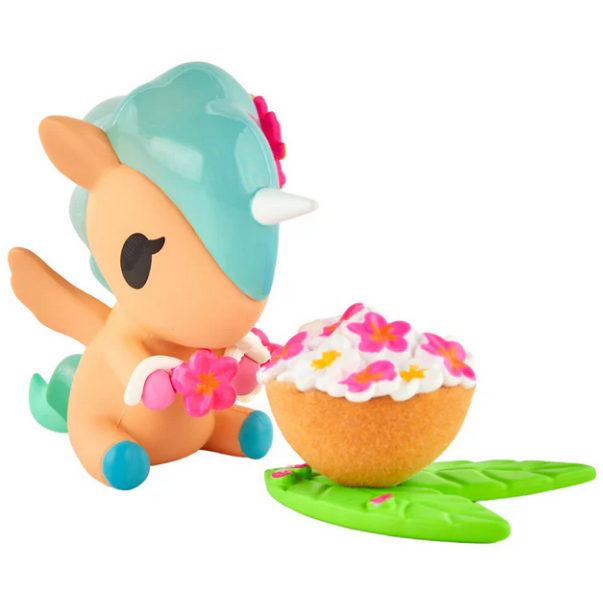 A small tokidoki Hawaiian Ohana Blind Box figurine with a blue mane and horn sits next to a flower-topped cupcake on green leaves, holding a pink flower garland—perfect for collectors and fans.