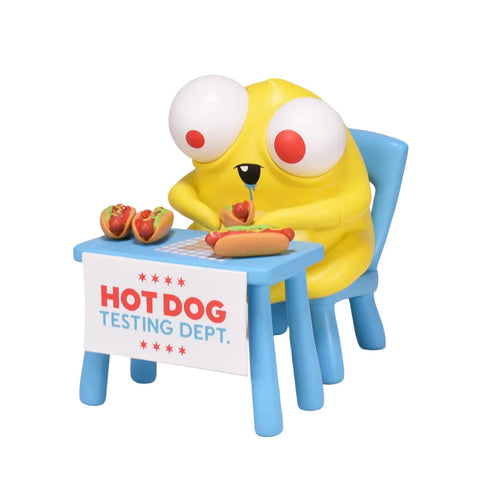 A yellow cartoon character with big eyes sits at a table labeled "Hot Dog Testing Dept." by Andrew Bell, carefully inspecting hot dogs on plates as part of the National Hot Dog Testing.