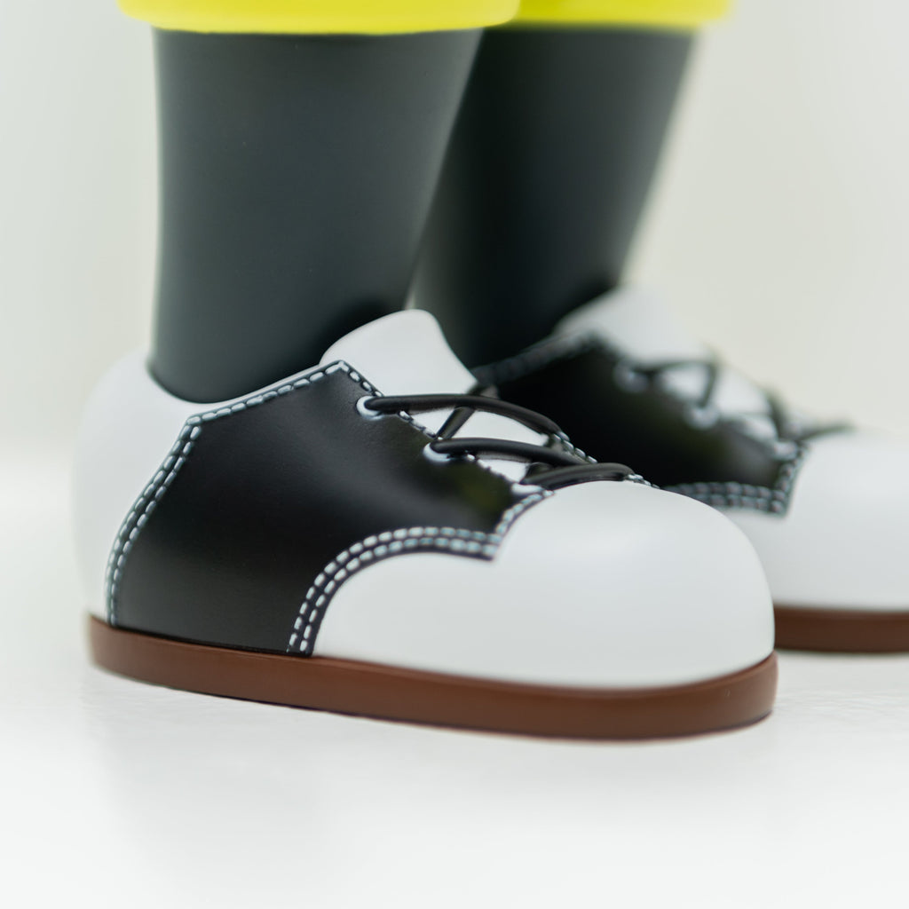 A close-up view of a pair of black and white lace-up toy shoes with brown soles, attached to a limited edition Hebru Brand vinyl figurine named Phibby — OG (Green), standing on a white surface.