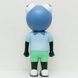 A figure named Phibby — Purple, featuring a frog-shaped head, a blue shirt, green shorts, and black limbs, stands facing away against a white background. This limited edition piece by Hebru Brand (US) beautifully captures the charm of Hebru Brantley's iconic characters.