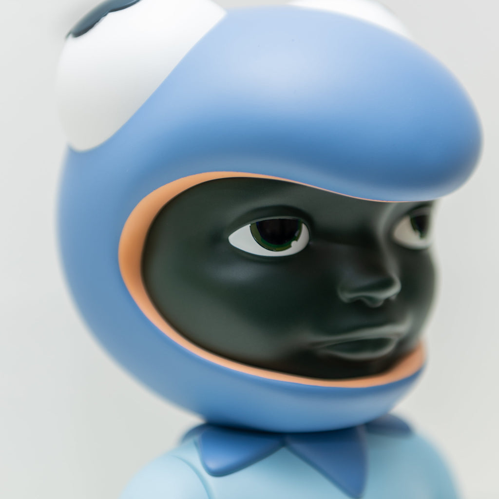 Close-up of the Phibby — Purple figurine by Hebru Brantley, showcasing dark skin and wearing a blue helmet that resembles a fish or creature with large eyes on top. The neutral expression complements the artist's iconic characters, signaling it might be part of a limited edition collection from Hebru Brand (US).