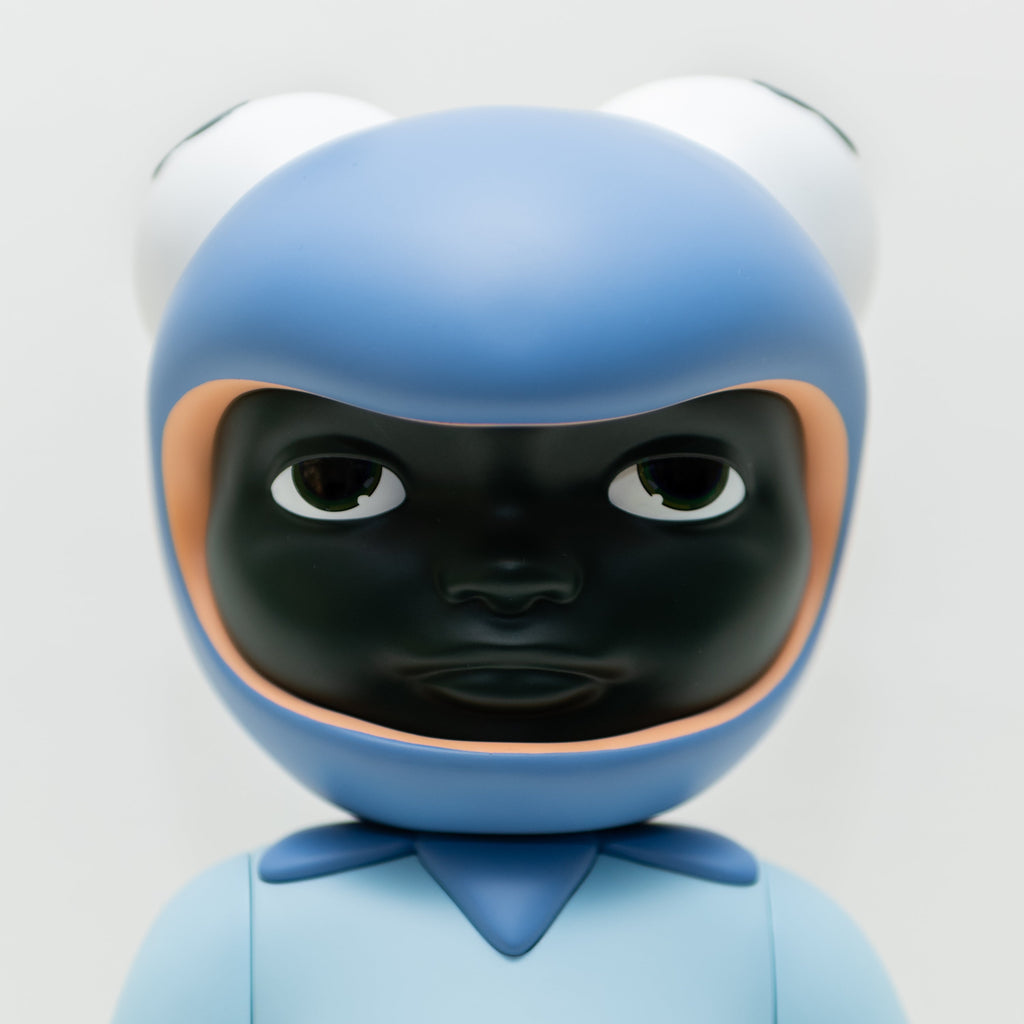 A close-up of Phibby — Purple, a limited edition figurine by Hebru Brand (US), featuring a dark face, large eyes, and a blue helmet resembling a frog's head with prominent eyes on top. The figurine’s body is also blue with a simplistic design, reminiscent of iconic characters created by Hebru Brantley.