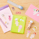 A desk with a pink notebook, a green notebook, a pink envelope, sticker packs of small dolls, a measuring tape, a pink paper clip, a pink glasses clip, and Sonny Angel dolls. There are also Dreams' water-resistant Sonny Angel Stickers — Volume 1 Blind Pack adding flair to the setup.