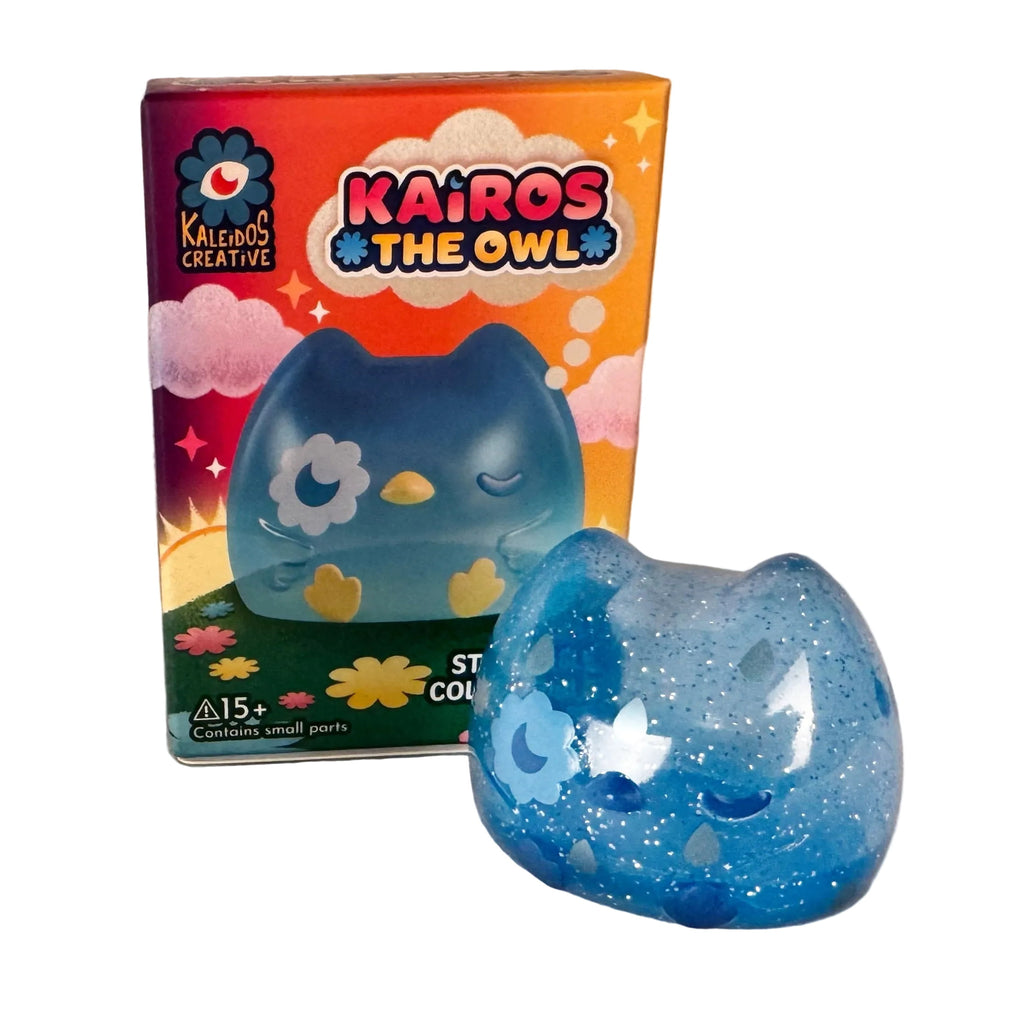 A blue, glittery owlet figurine from the Kairos the Owl Blind Box by Kaleidos Creative sits before its colorful packaging; this collectible is a random figure recommended for ages 15+.