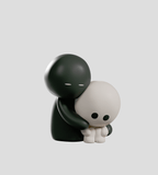 A matte black Meyanmi Shadow Blind Box figure by Kaleidos Creative hugs a smaller, matte white figure sitting with knees up, both displayed against a plain gray background.