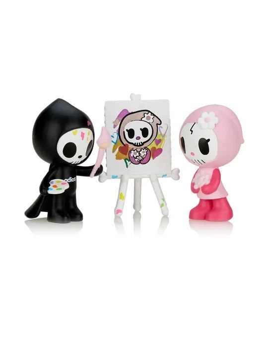 Two cartoon skeleton figures, one in black painting clothes holding a palette and brush, the other in pink, stand by an easel displaying a painted skeleton artwork. This whimsical scene is part of the