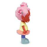 A side view of Rotofugi’s Binky — OG, a limited edition art toy inspired by Bianca Pastel, featuring a Black girl resin figure with pink hair in buns, yellow shirt, pink skirt, and blue boots standing upright against a white background.