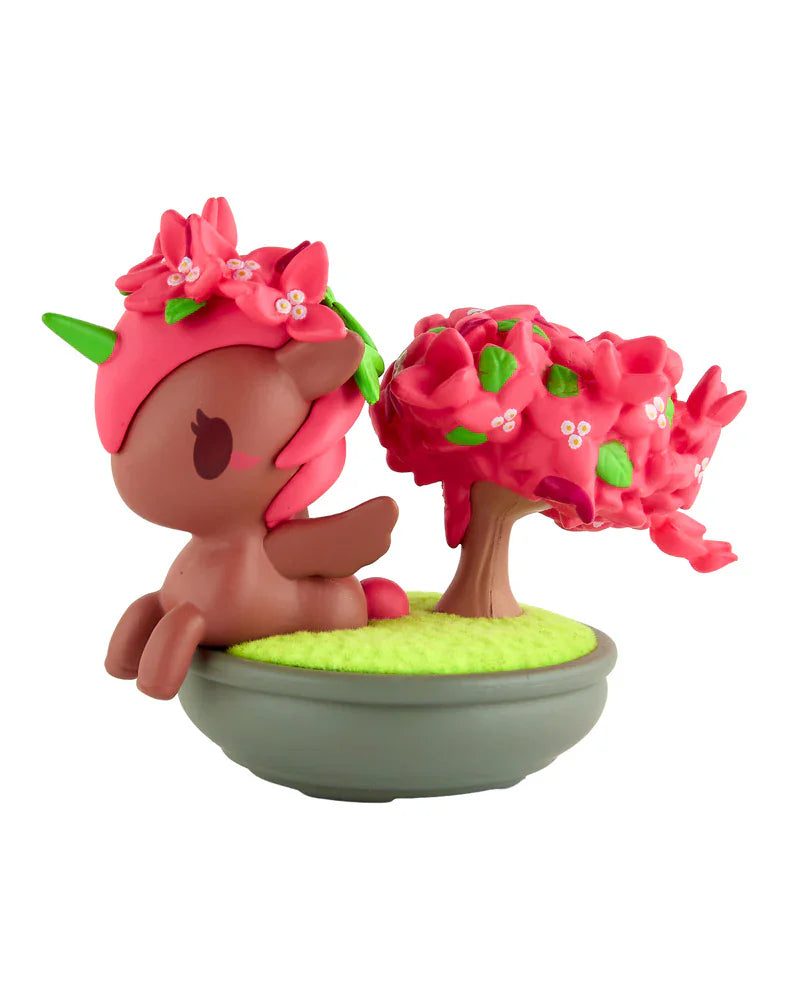 A brown unicorn with a green horn and pink flower mane from the tokidoki Botanical Bonsai Unicorno Blind Box sits beside a matching pink flowering tree in a round gray pot—perfect for miniature garden displays.