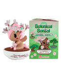 A small unicorn figurine with pink hair, antlers, and flowers sits in a bonsai dish next to a green Tokidoki Botanical Bonsai Unicorno Blind Box—ideal for tokidoki fans and collectors of whimsical garden-themed miniatures.