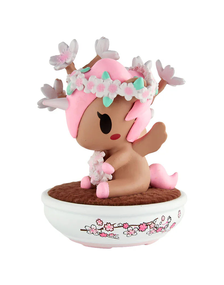 A small pink and brown deer-like figurine with wings and a flower crown from the tokidoki Botanical Bonsai Unicorno Blind Box sits in a white bowl with cherry blossom designs on a brown surface.