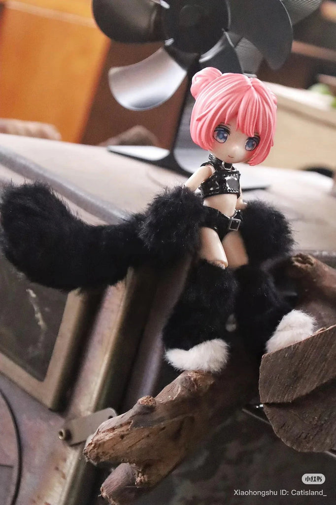 A small BIJIDI Cat Island — Black Cat Or Candy Cat? Blind Box doll with pink hair, blue eyes, and a black-and-white furry outfit sits on a branch indoors with a blurred fan in the background.