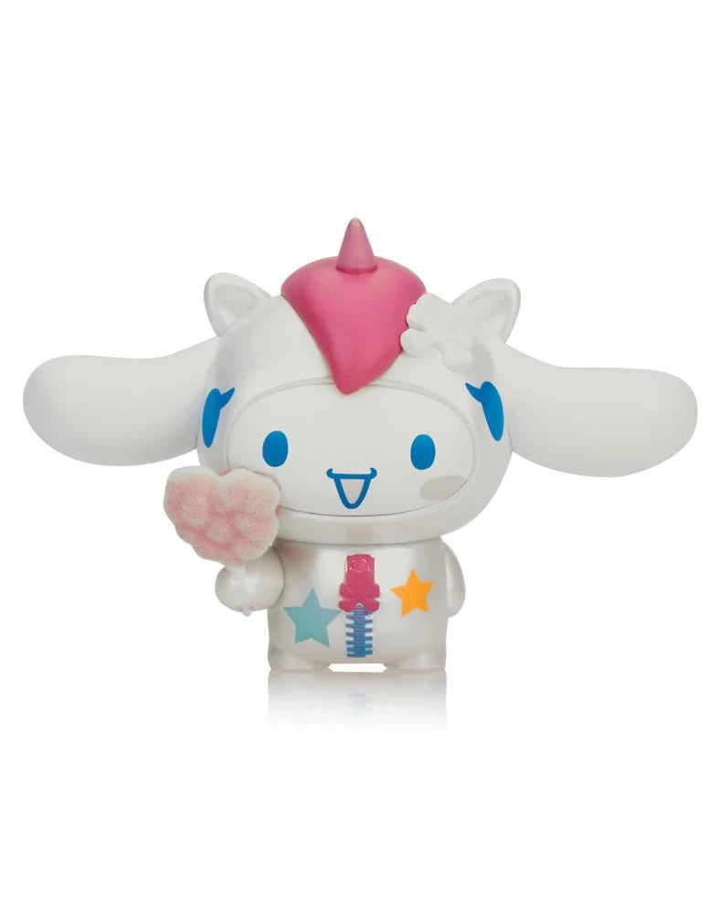 A white cartoon figure with blue accents, a pink unicorn horn, and long ears holds a pink paw object. It features stars and a flower on its head and is part of the Tokidoki x Cinnamoroll Sweet Treats Blind Box series by Tokidoki.