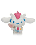 A white cartoon figure with blue accents, a pink unicorn horn, and long ears holds a pink paw object. It features stars and a flower on its head and is part of the Tokidoki x Cinnamoroll Sweet Treats Blind Box series by Tokidoki.