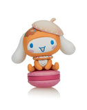 A cute orange-and-white Tokidoki x Cinnamoroll figure with floppy ears, a donut hat, and blue eyes sits on a pink macaron—part of the Tokidoki Sweet Treats Blind Box series.