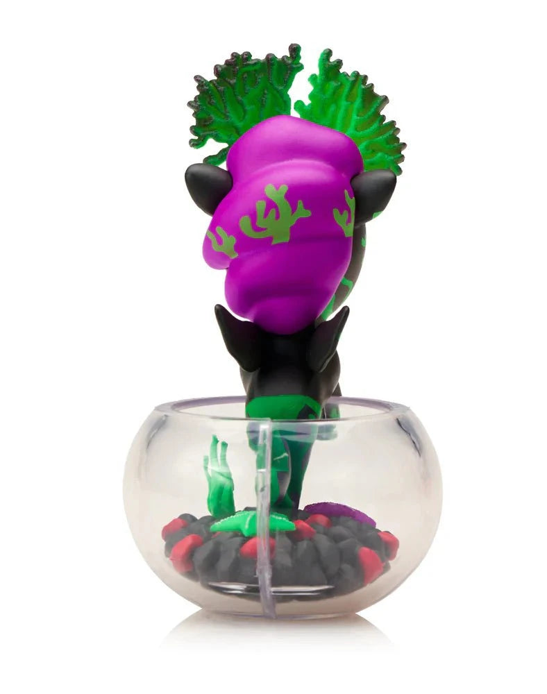 The Tokidoki Neon Reef Mermicorno - Deep Star Coral Special Edition is a small black and green figurine with purple hair, leaf details, and sits upright in a clear round container filled with glow-in-the-dark black and red pebbles.