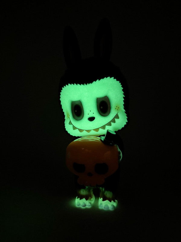 Labubu - Halloween by How2Work (HK) is a glow-in-the-dark figurine featuring rabbit ears, a wide smile, and dark eyes, holding an orange skull—an essential collectible for toy enthusiasts.