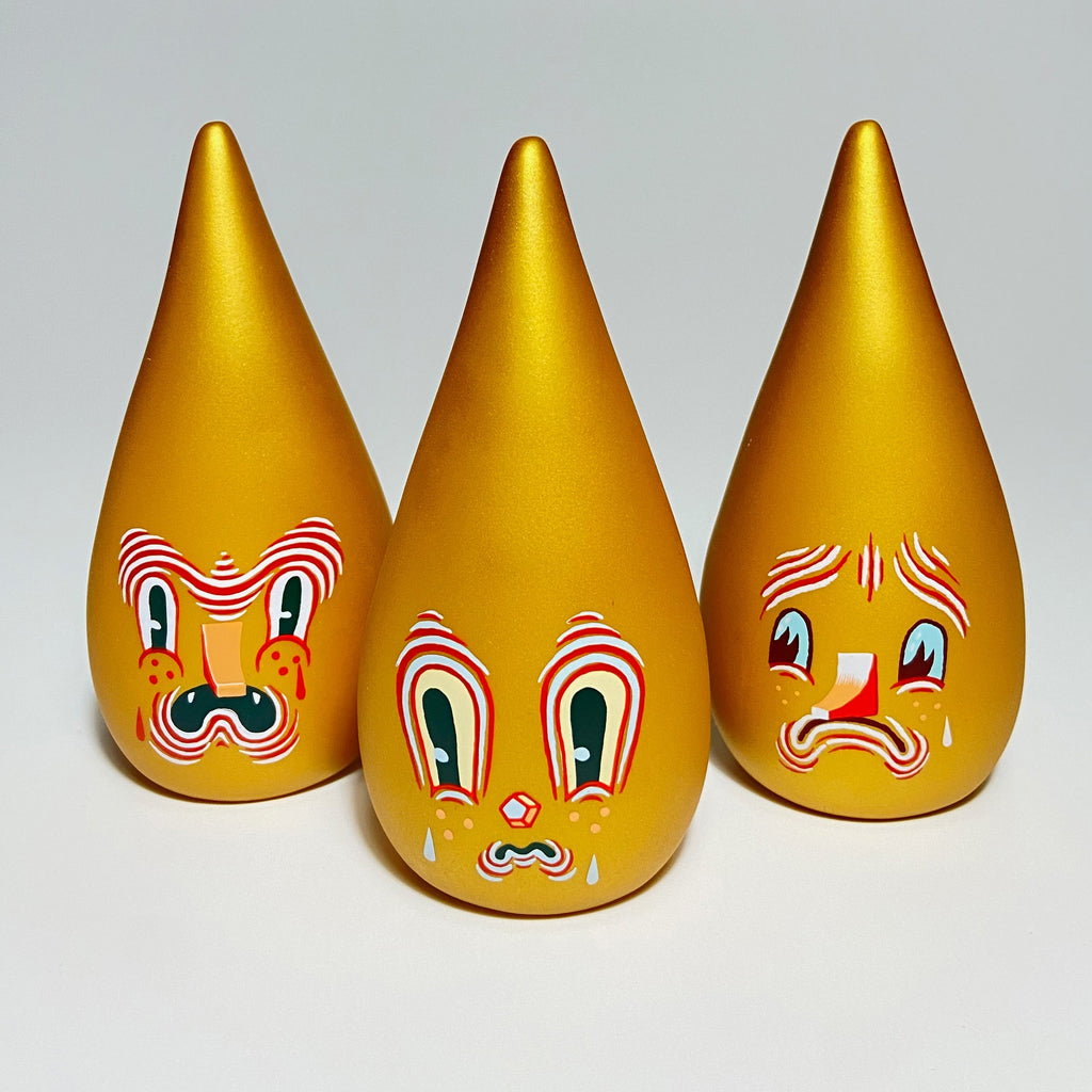 Three golden, teardrop-shaped figurines from the Mini Driplets — Primo & Plebe Blind Box by Squibbles Ink + Rotofugi (US) collection with expressive, painted faces depicting different emotions: left one concerned, middle one surprised, and right one sad.