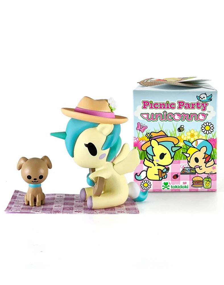 A unicorn figurine with a hat and wings sits on a picnic blanket beside a small dog, featuring collectible figures from the Tokidoki Picnic Party Unicorno Blind Box by tokidoki (IT), with its colorful packaging shown behind them.