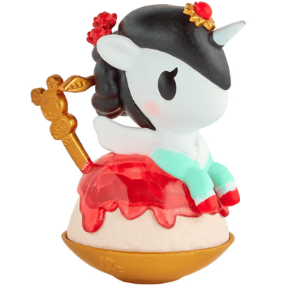 A small tokidoki Shaved Ice Dreams Unicorno Blind Box figurine, featuring black hair, a jeweled horn, a red dress, aqua legs, and a golden teacup-style handle, sits atop a round cream and gold base. Perfect for collectors and fans who enjoy the fun surprise of a blind box toy.