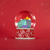 The Dumpster Fire Snow Globe by 100% Soft has a green dumpster with yellow and purple trash on a red base with white line art, making quirky seasonal desk decor against a red background with white specks.