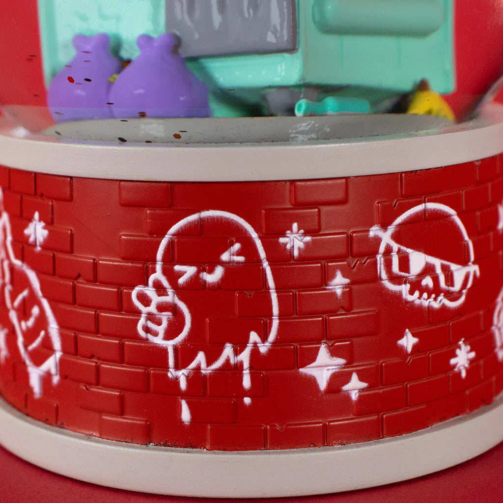Close-up of the 100% Soft Dumpster Fire Snow Globe shows a red plastic surface with white graffiti-style ghost, skull, and star drawings on a brick-pattern background—an edgy accent for your seasonal desk or snow globe collection.