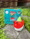 A Rotofugi Sushi Snails Blind Box collectible, featuring a toy snail styled like sushi on a stone with green plants, is shown with its colorful packaging in the background—ideal for fans of fun art toys.