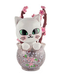 A ceramic white cat with big green eyes sits in a floral vase amid pink cherry blossoms, evoking the charm of cherry blossom season and celebrating the Tokidoki Cherry Blossom Cats - Single Blind Box by Tokidoki.
