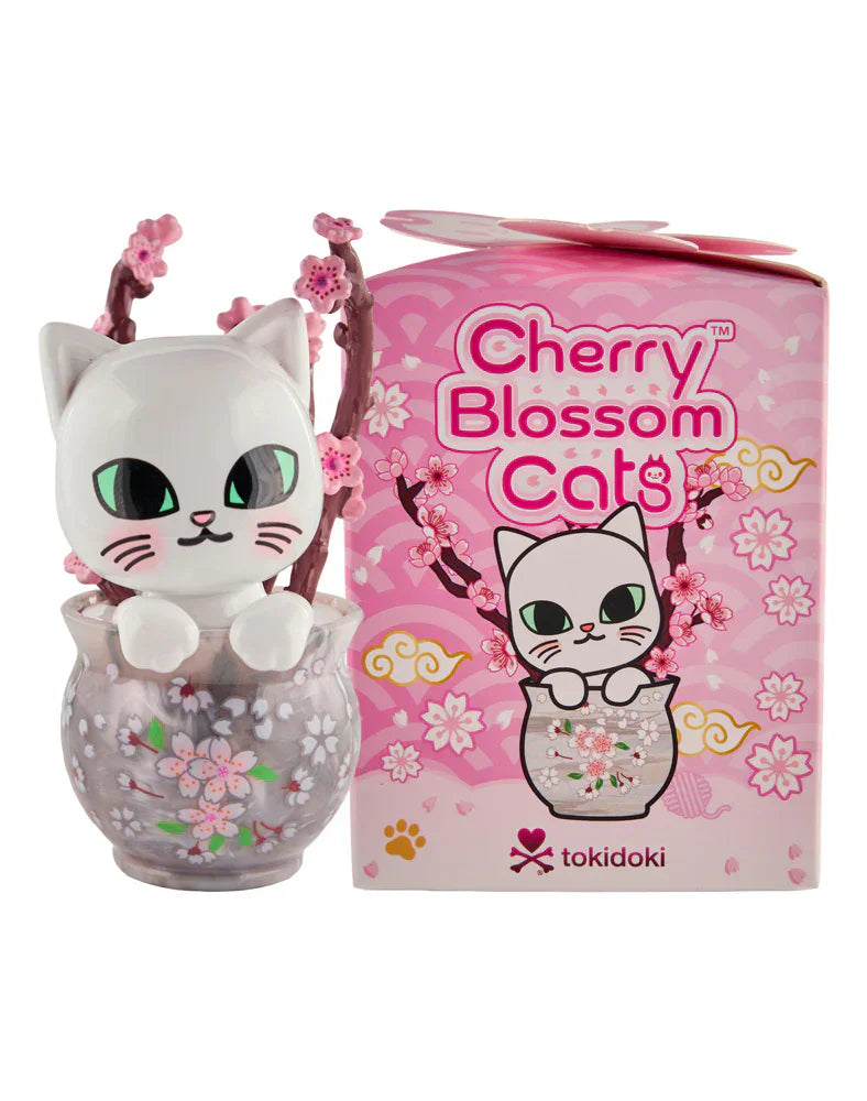 A white cat figurine with green eyes and cherry blossom accents sits in a clear bowl beside a Tokidoki Cherry Blossom Cats- Single Blind Box from Tokidoki, featuring artwork inspired by cherry blossom season.