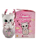 A white cat figurine with green eyes and cherry blossom accents sits in a clear bowl beside a Tokidoki Cherry Blossom Cats- Single Blind Box from Tokidoki, featuring artwork inspired by cherry blossom season.