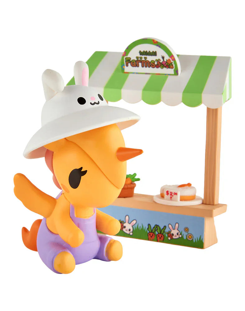 A tokidoki Farmers Market Unicorno Blind Box figure—a small orange dragon in a white bunny hat and purple outfit—sits before a green-striped stand with cake and plant accents, perfect for tokidoki fans and collectors.
