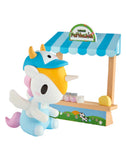 A tokidoki Farmers Market Unicorno Blind Box figure, styled as a cow, sits by a toy farm stand with a blue-striped roof and tiny jars and cheese, inspired by the collectible series from tokidoki.