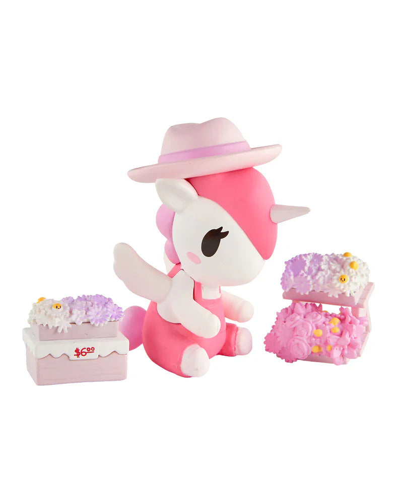 A pink and white tokidoki Unicorno figure with a hat is displayed between two tokidoki Farmers Market Unicorno Blind Boxes, each featuring pink and purple floral designs.