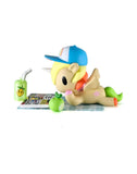 A Tokidoki Picnic Party Unicorno Blind Box figure by tokidoki (IT) lounges with a blue cap, reading on a mat beside an apple and juice box—a delightful collectible for fans.