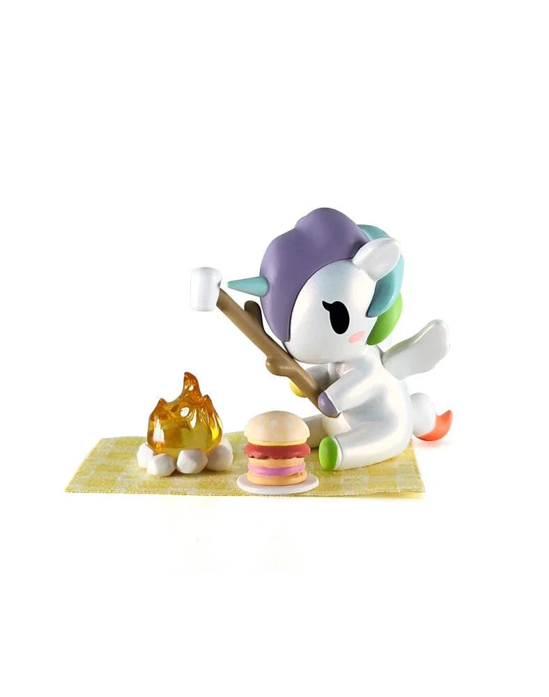 A Tokidoki Picnic Party Unicorno Blind Box figure from tokidoki (IT) roasts a marshmallow by a campfire with a sandwich and yellow checkered mat, making it perfect for whimsical displays.