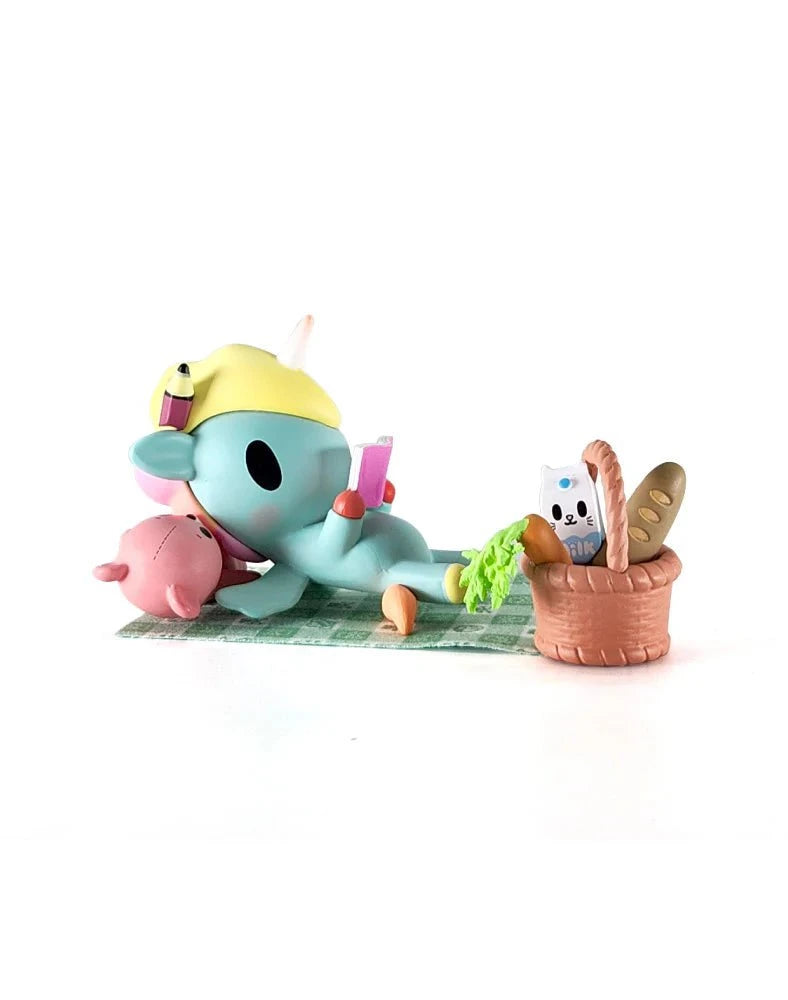 A blue cartoon animal in a yellow hat lounges on a mat with a pink animal at their tokidoki (IT) Picnic Party Unicorno, holding a drink beside a picnic basket with bread and a cat-face carton. Tokidoki Picnic Party Unicorno Blind Box.
