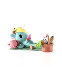 A blue cartoon animal in a yellow hat lounges on a mat with a pink animal at their tokidoki (IT) Picnic Party Unicorno, holding a drink beside a picnic basket with bread and a cat-face carton. Tokidoki Picnic Party Unicorno Blind Box.