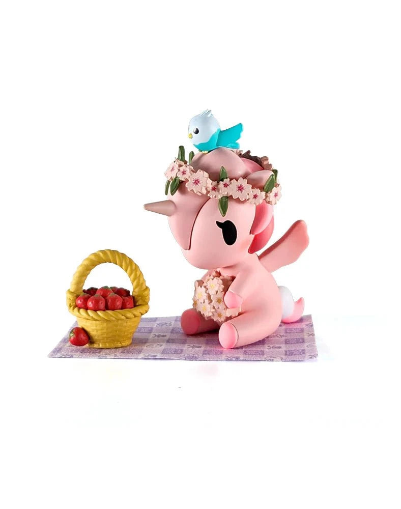 The Tokidoki Picnic Party Unicorno Blind Box by tokidoki (IT) features a pink Unicorno with a flower crown and bird, seated on a checkered mat beside a basket of red apples—a delightful collectible figure.