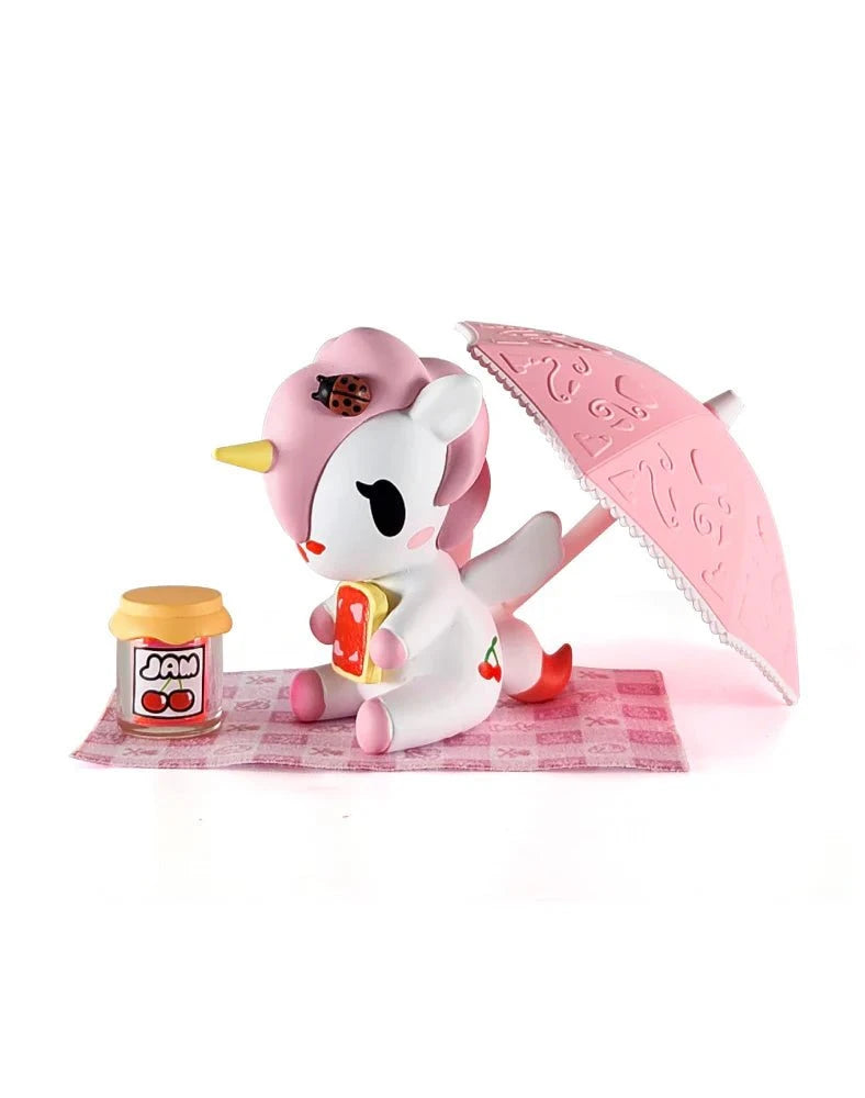 A Tokidoki Picnic Party Unicorno Blind Box figurine by tokidoki (IT) sits on a pink checkered mat holding toast, with a
