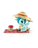 A small blue cartoon animal in a wide-brimmed hat sits on a red-checked picnic blanket, holding a sandwich by a