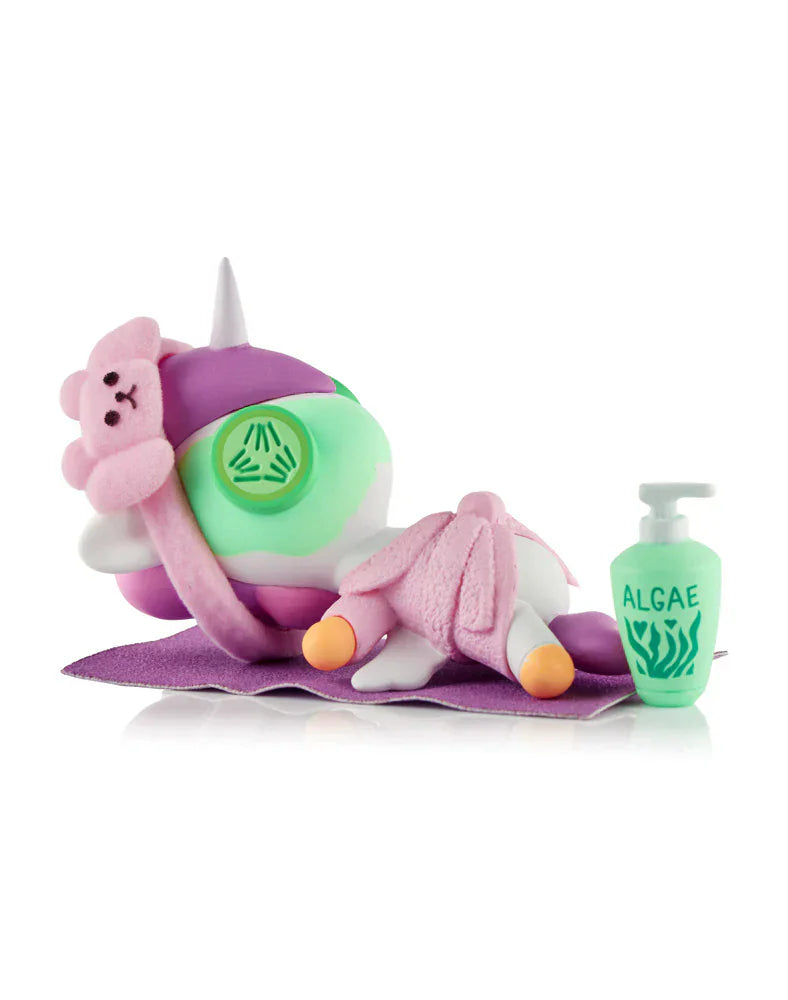 A Tokidoki Spa Day Unicorno Blind Box figurine lies face down on a purple mat with a bear eye mask, towel, and