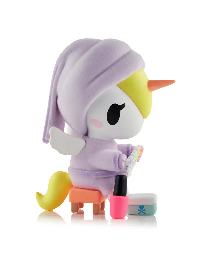 A Tokidoki Spa Day Unicorno Blind Box figurine features a kawaii Unicorno in a lavender robe and cap, seated on a stool with a nail polish palette, nail polish bottle, and small container beside it.